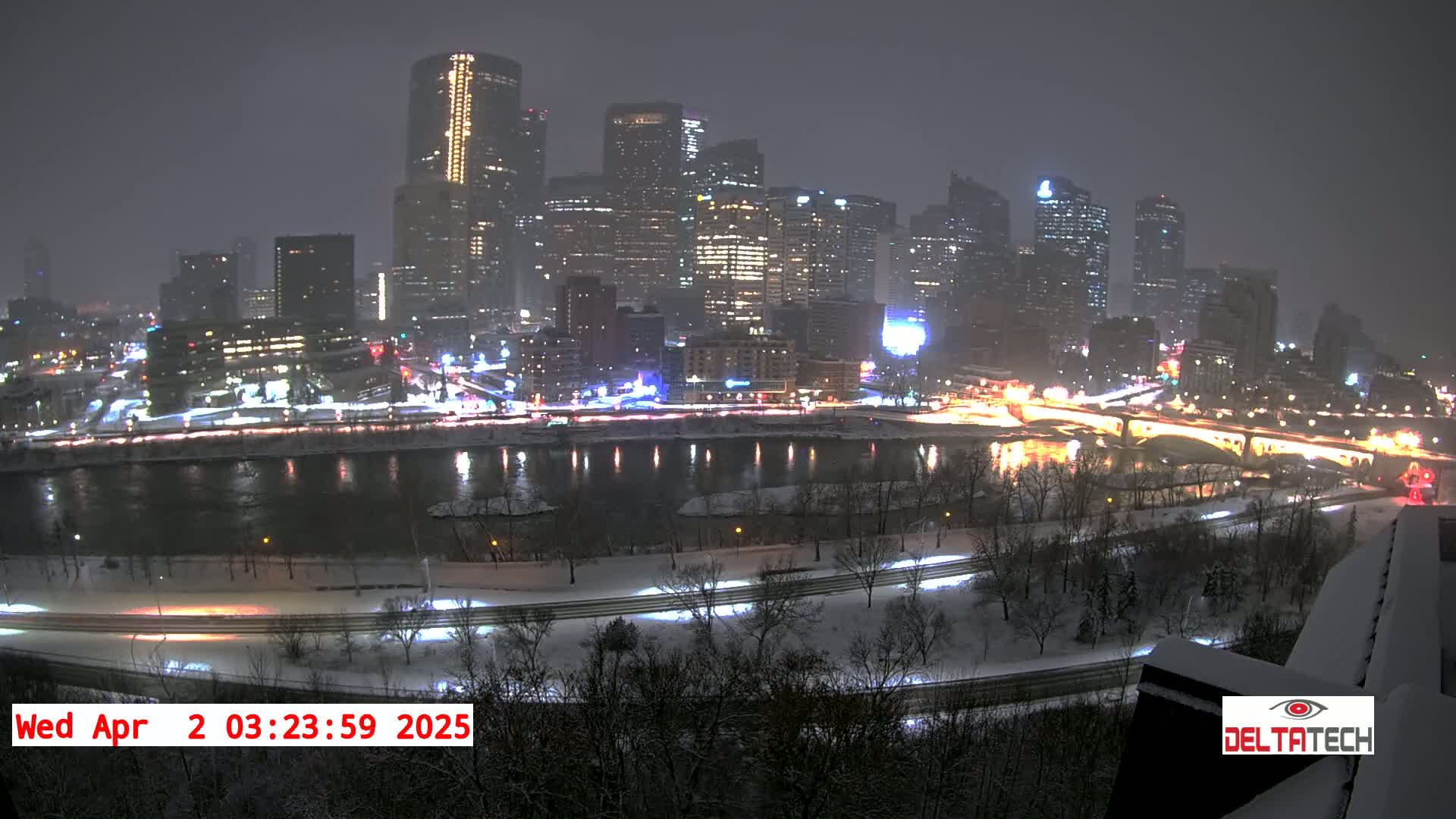 A snow-covered city skyline at night, illuminated by brightly lit skyscrapers and streetlights, is reflected in a calm river.