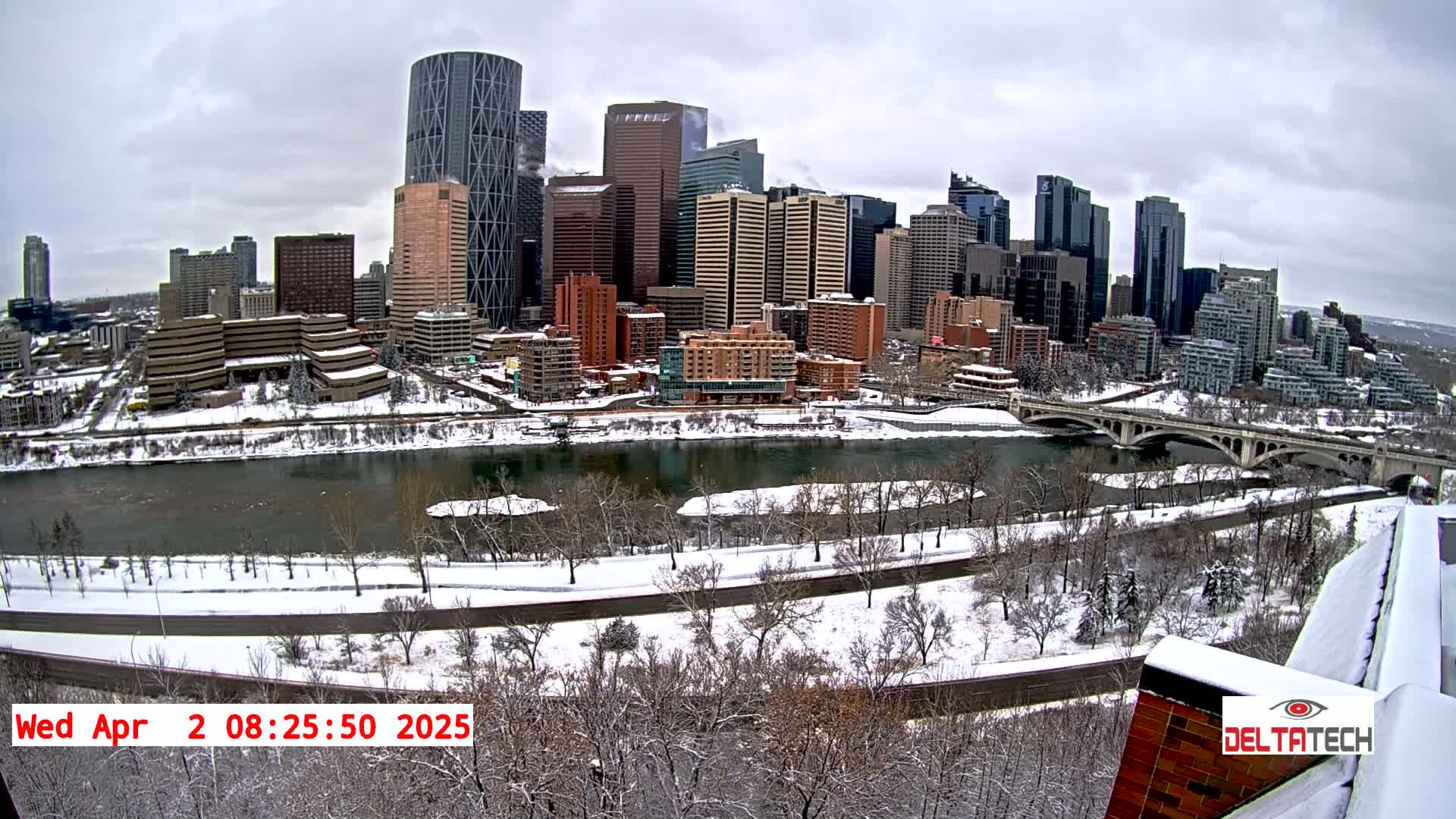 A snow-covered city skyline with a river and bridge is seen on an overcast day.