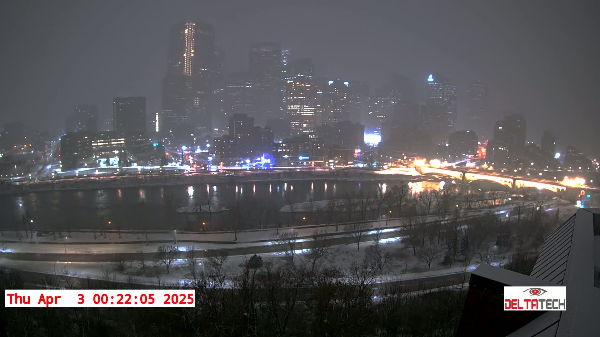 A snow-covered city skyline is illuminated at night, reflected in a dark river below.