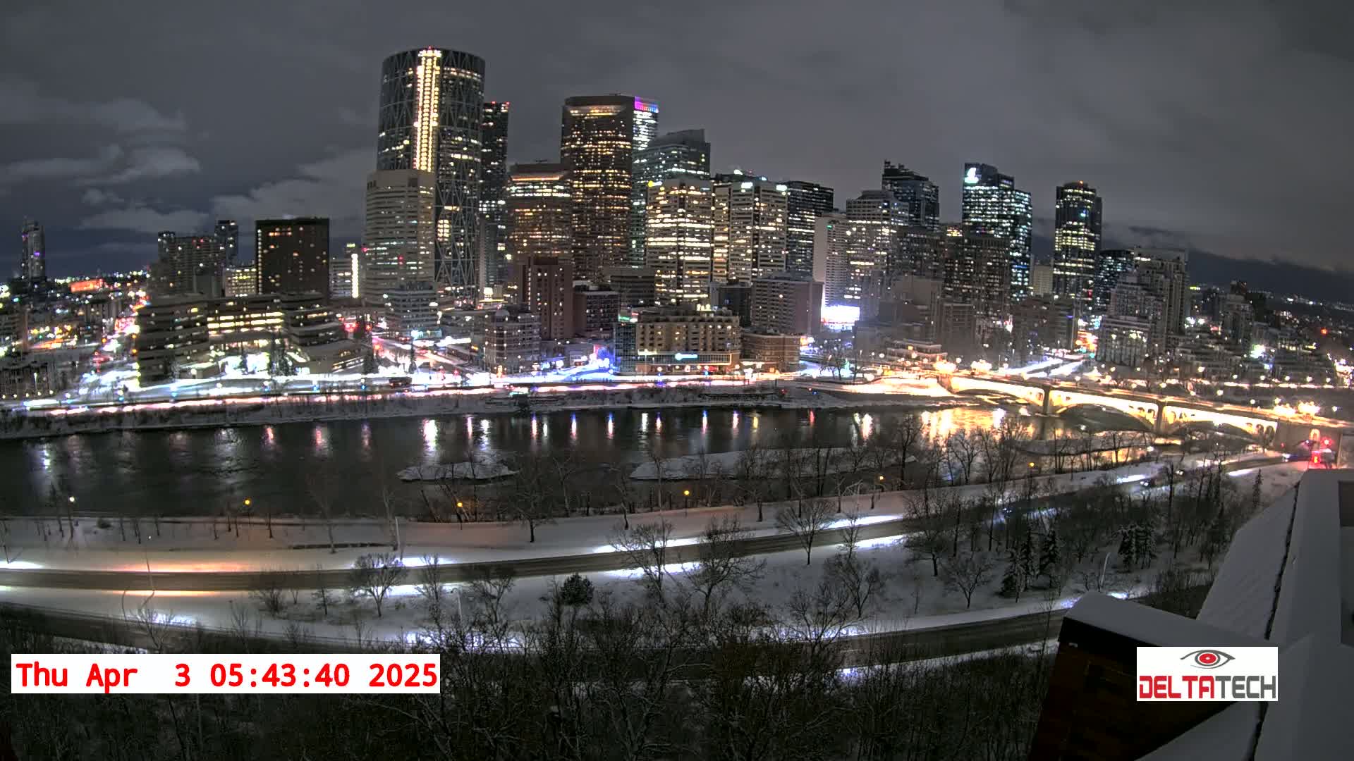 A snow-covered city skyline at night is illuminated by bright lights, reflected in a dark river flowing below.