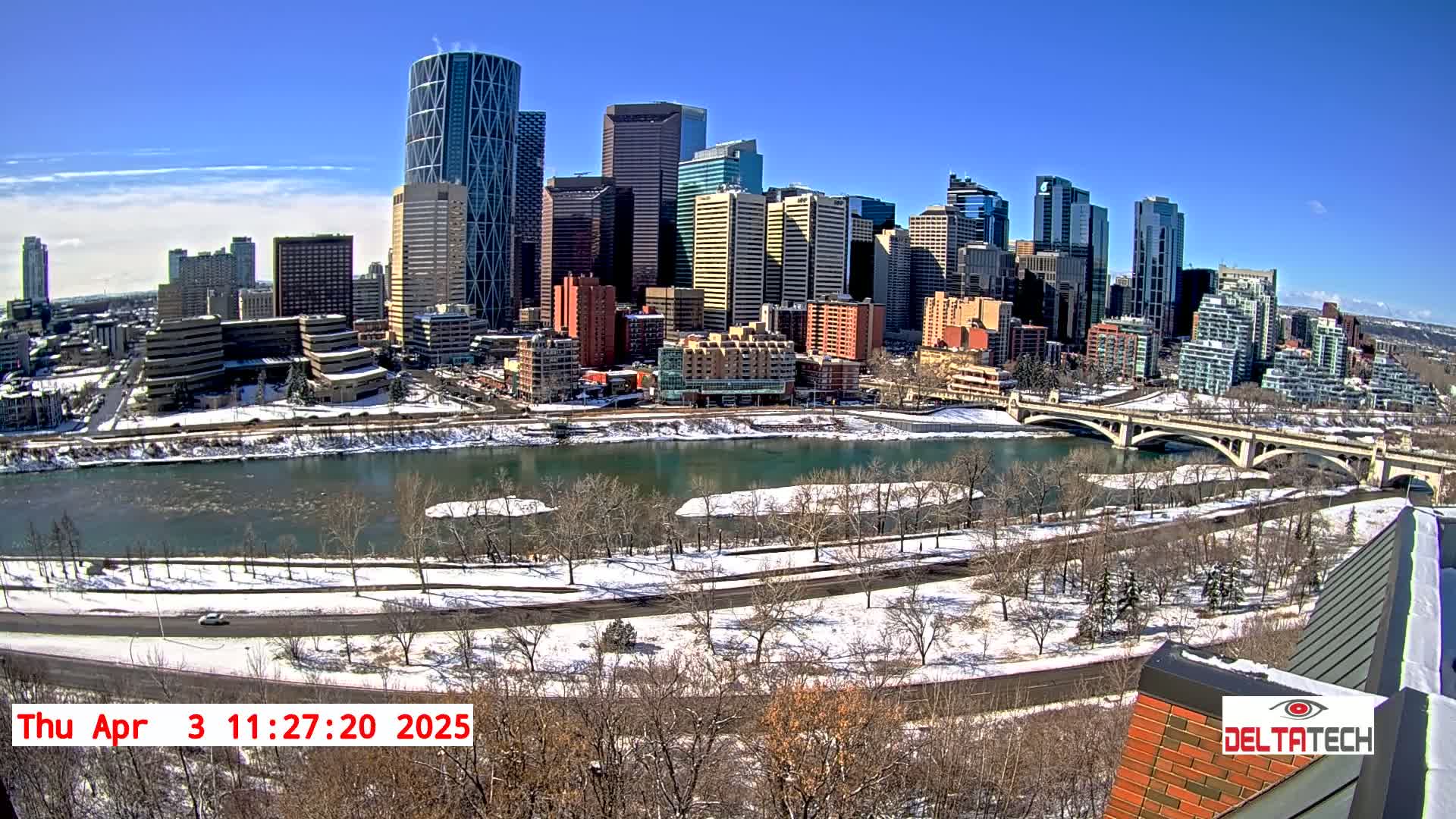 A city skyline with numerous skyscrapers is visible across a snow-covered river on a sunny day.