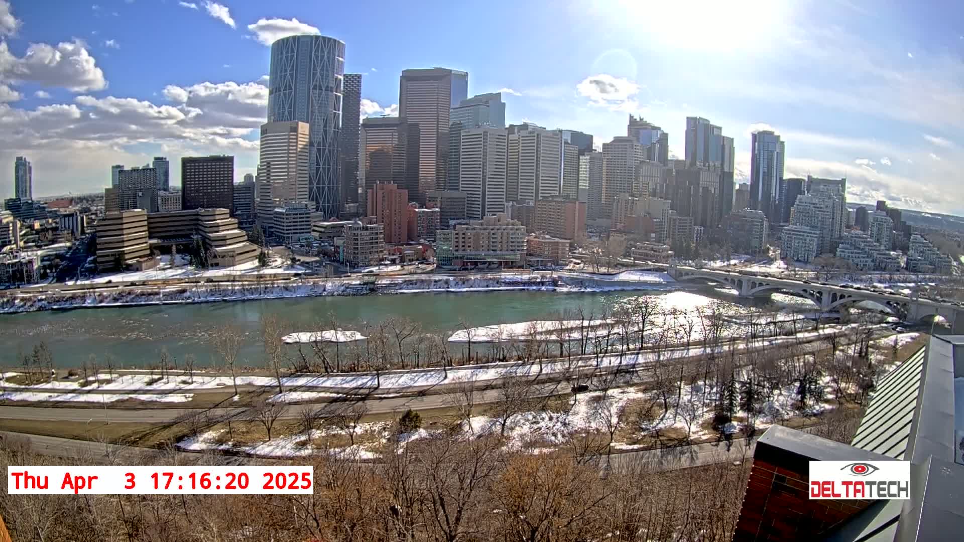 A sunny day reveals a city skyline across a partially frozen river, with snow on the ground and bare trees lining the riverbanks.