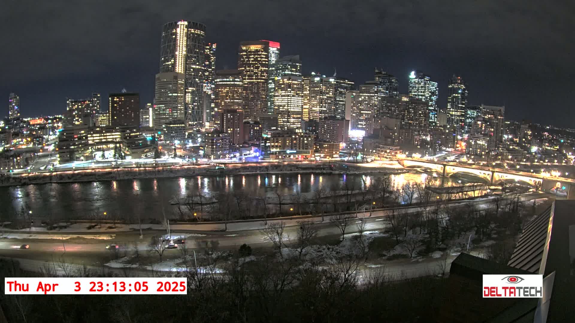A nighttime view of a city skyline illuminated along a river with snow on the ground and bare trees.
