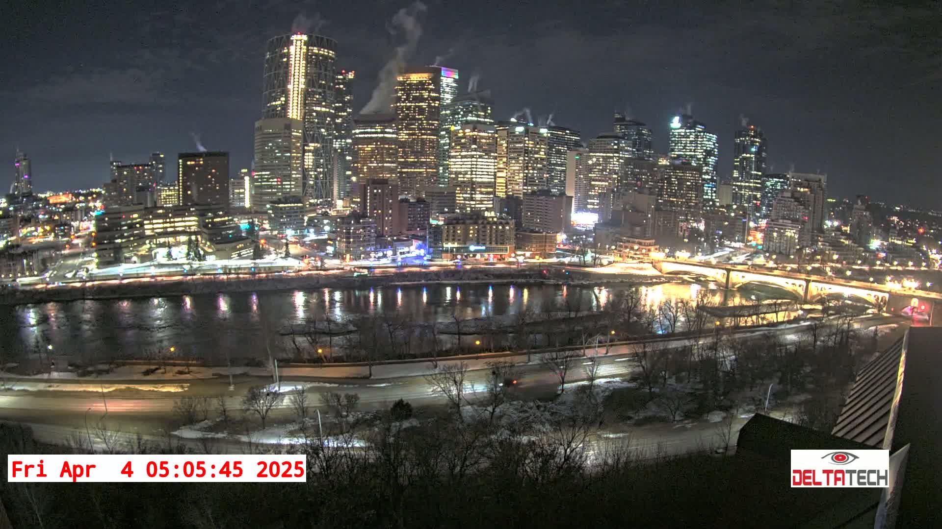 A nighttime cityscape illuminated with city lights, reflecting on a river partially covered with ice and snow.