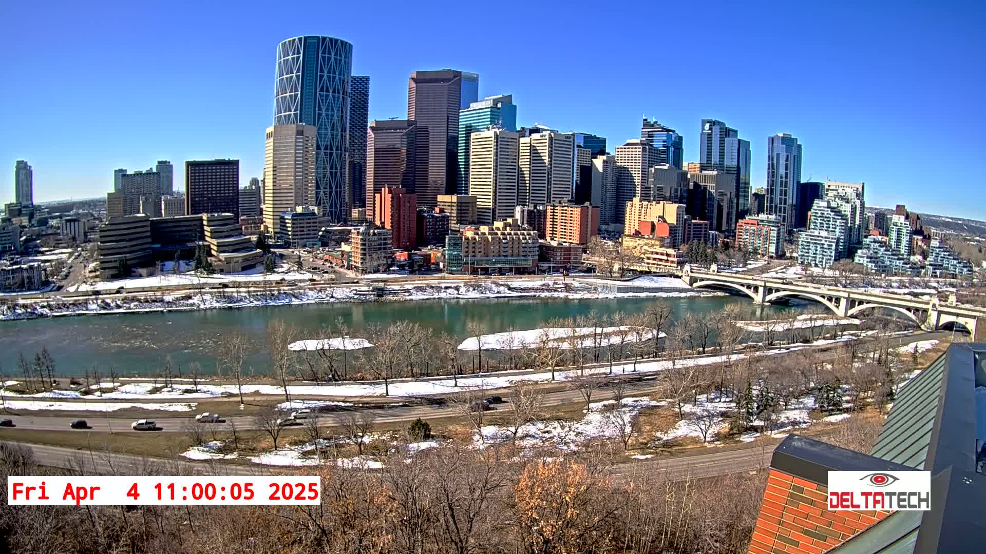 A city skyline with many skyscrapers is seen across a river on a sunny day with patches of snow on the ground and along the riverbank.