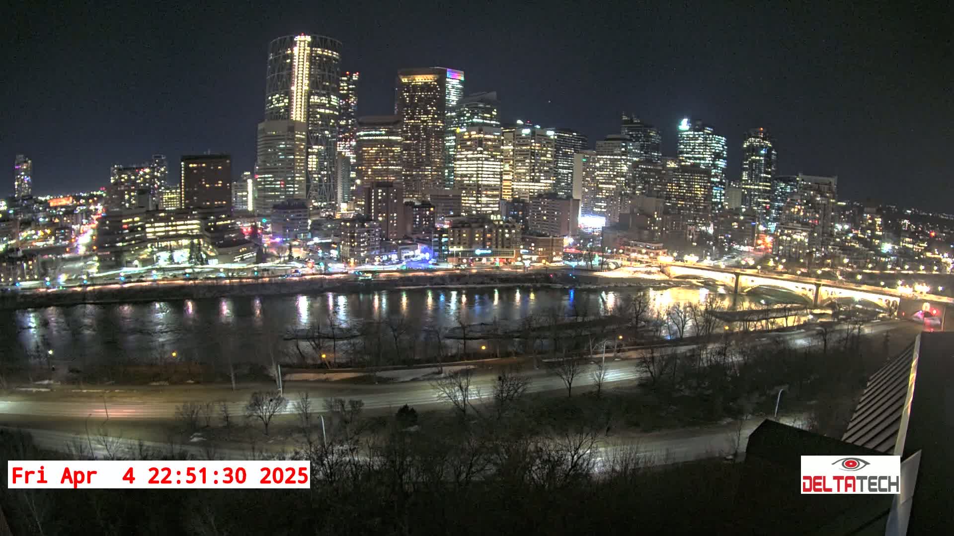 A nighttime view of a city skyline illuminated across a calm river, with patches of snow visible on the ground.
