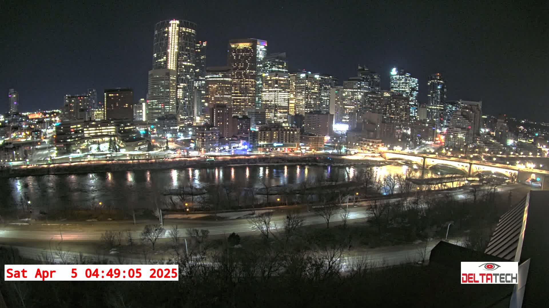 A nighttime view of a city skyline illuminated with lights, reflected in a calm river bordered by roads and bare trees with some snow on the ground.