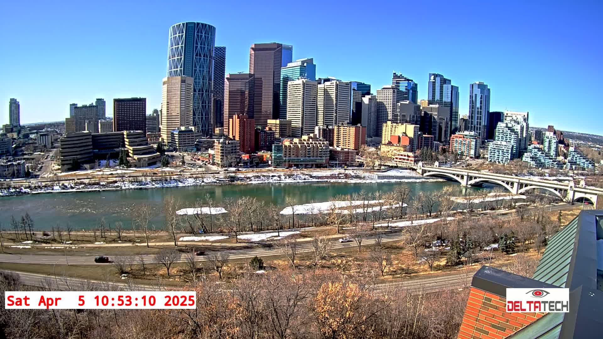 A city skyline with numerous skyscrapers and high-rise buildings is seen across a river with patches of ice and snow along its banks under a clear, sunny sky.