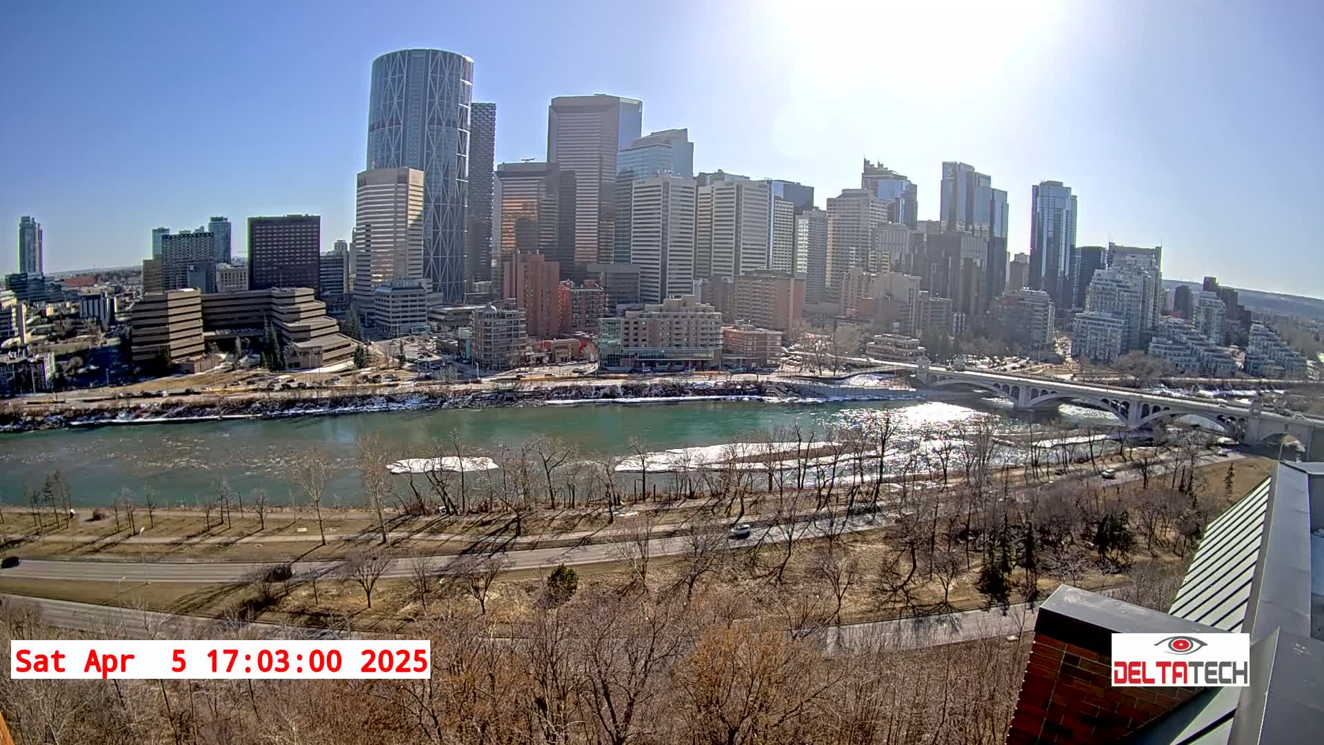 A sunny cityscape with a river partially frozen over, viewed from a high vantage point.