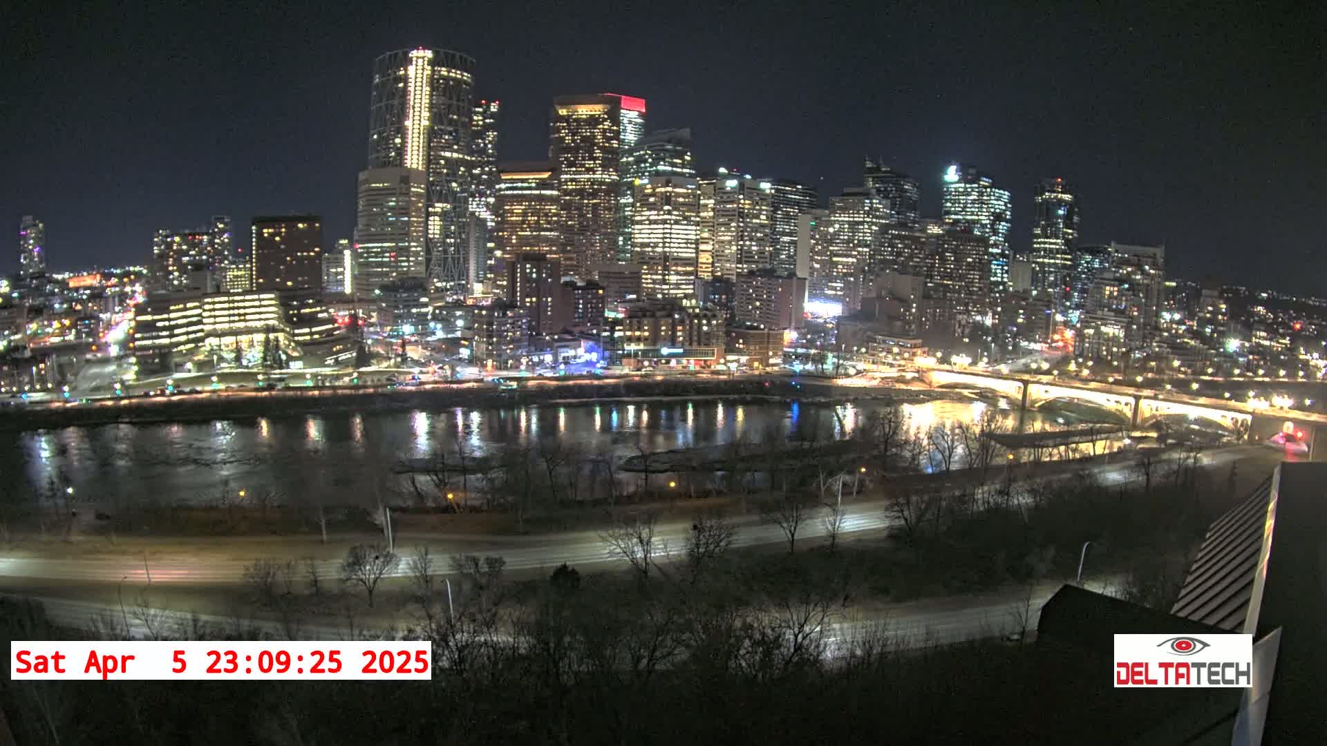 A brightly lit city skyline at night is reflected in a dark river, with a road and bare trees in the foreground under a clear sky.