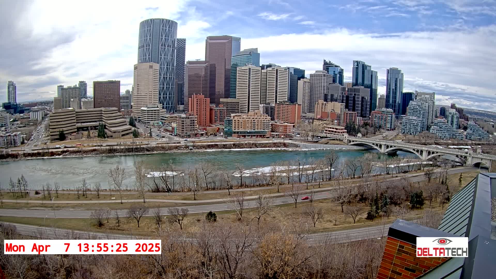 A city skyline with numerous skyscrapers is situated across a river with partially frozen water, under a partly cloudy sky.