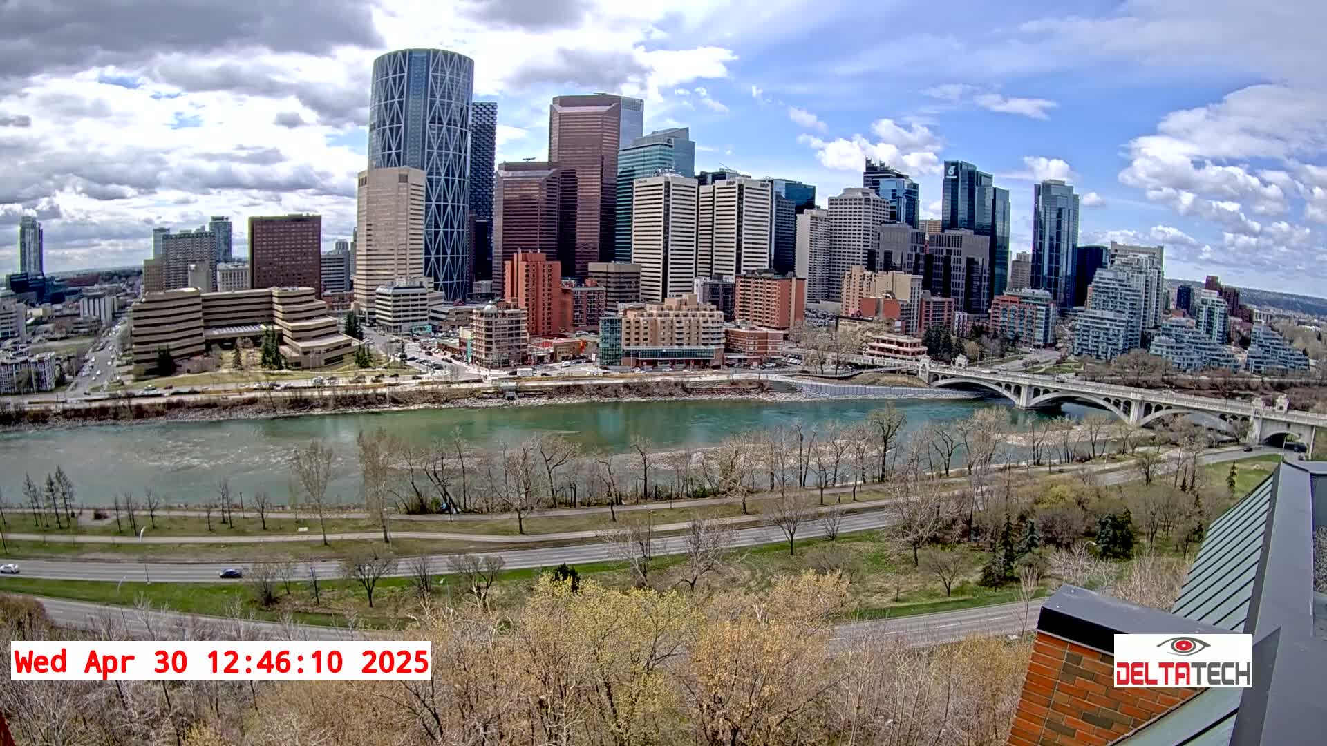 A city skyline with numerous skyscrapers sits across a wide river under partly cloudy skies, with bare trees lining the riverbank and a bridge spanning the water.