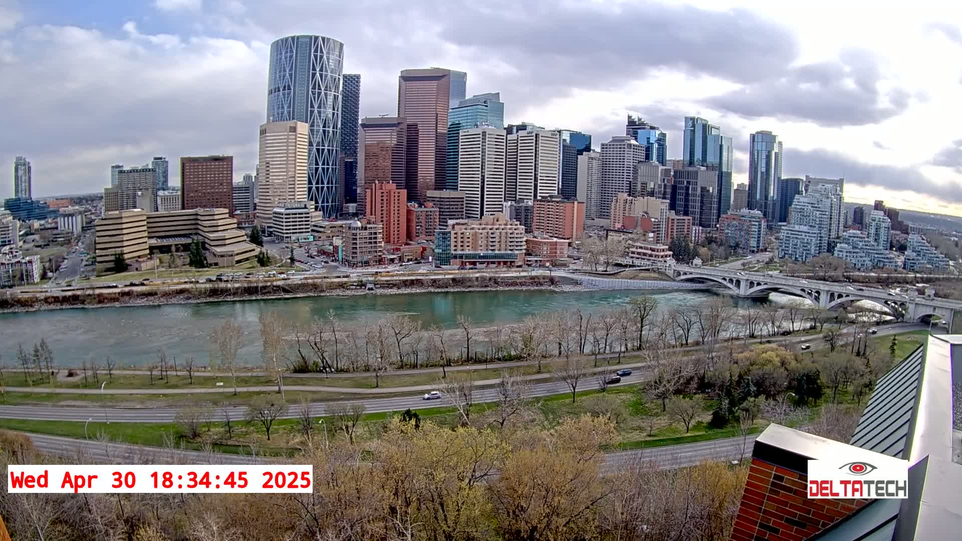 A city skyline with many tall buildings is seen across a river with a bridge under partly cloudy skies.