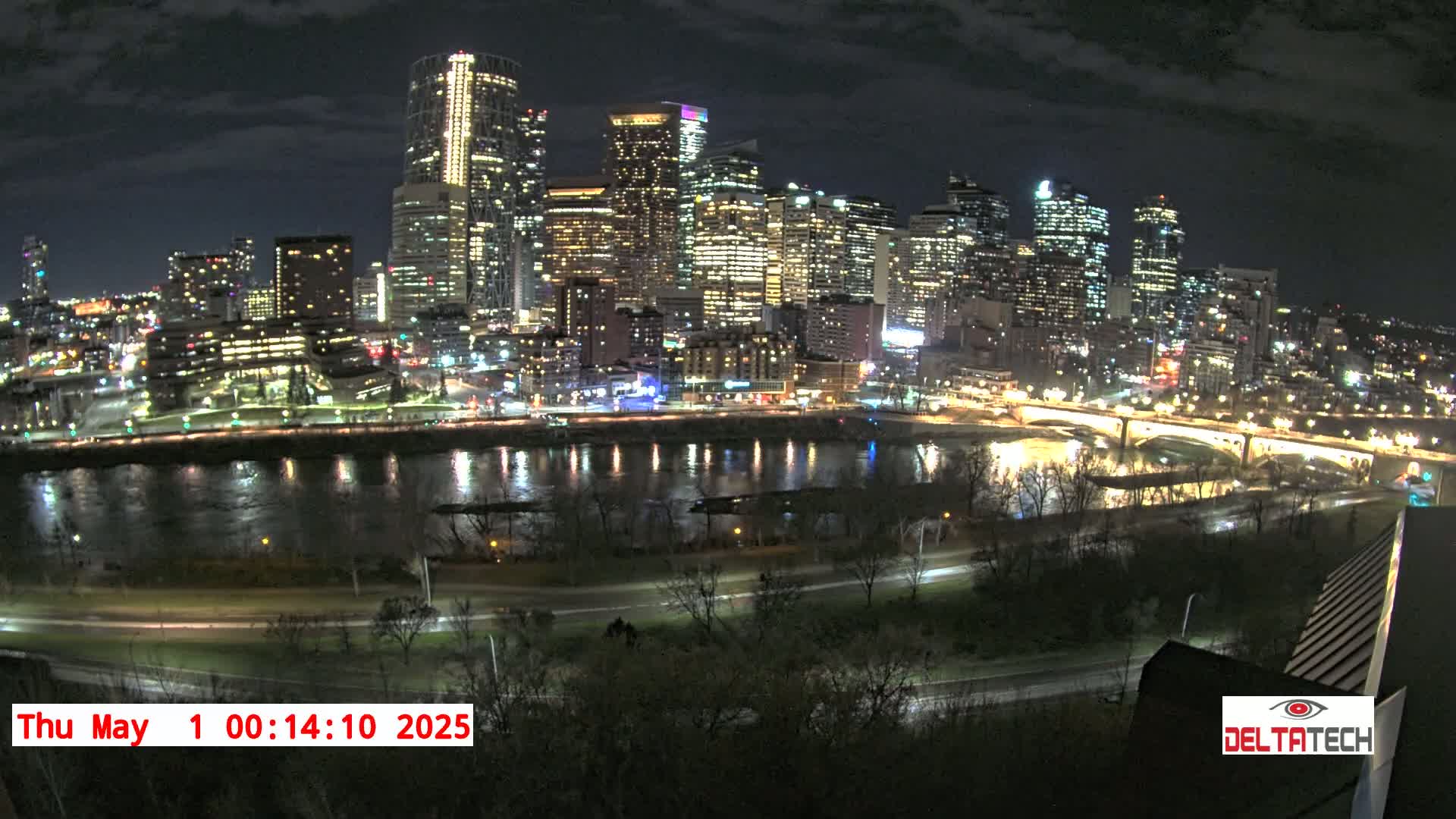 A nighttime view of a city skyline illuminated with lights, reflected in a calm river below, under a partly cloudy sky.