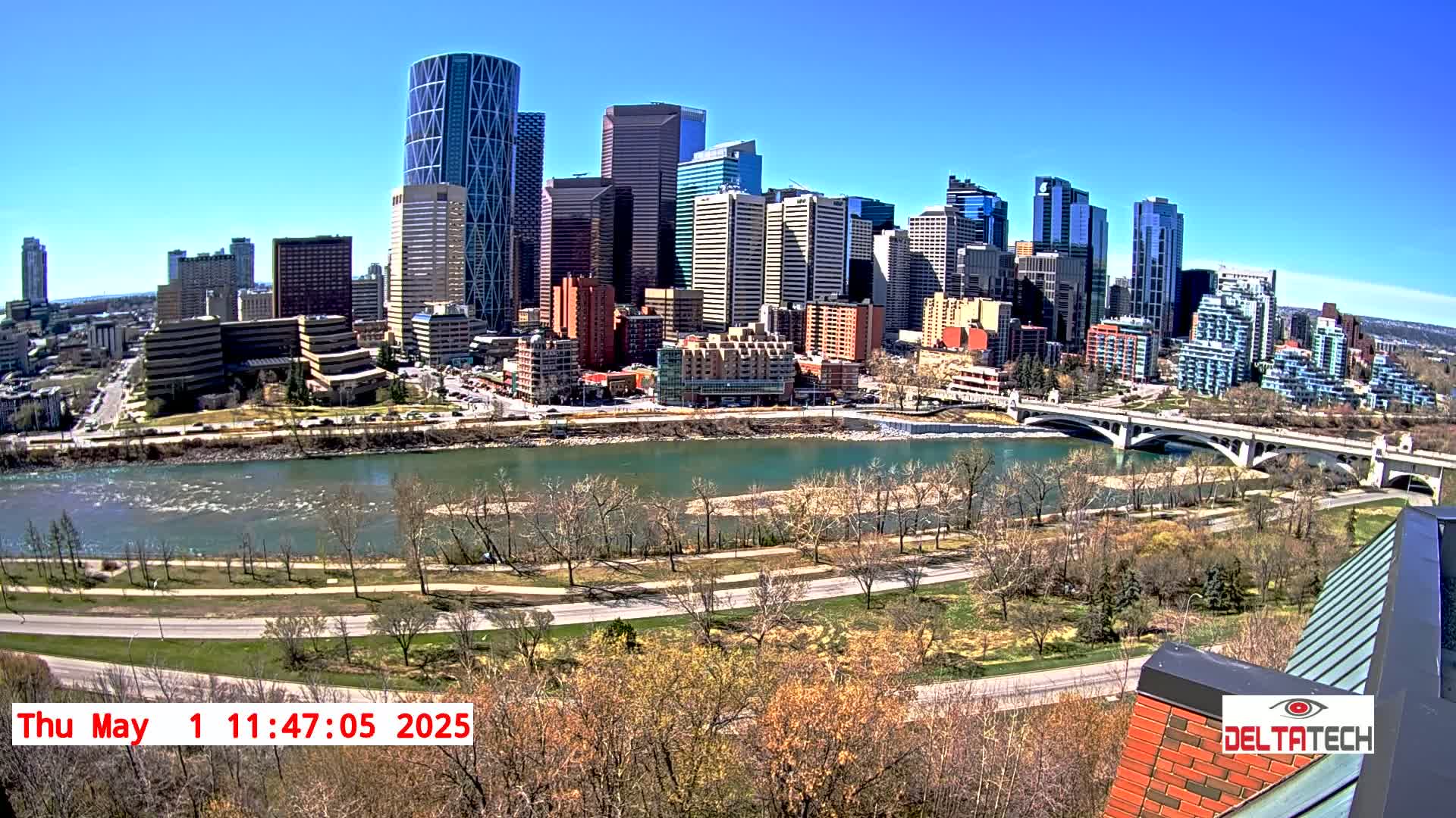 A cityscape with numerous skyscrapers is situated across a river on a sunny day, with a bridge connecting the city to the other side.