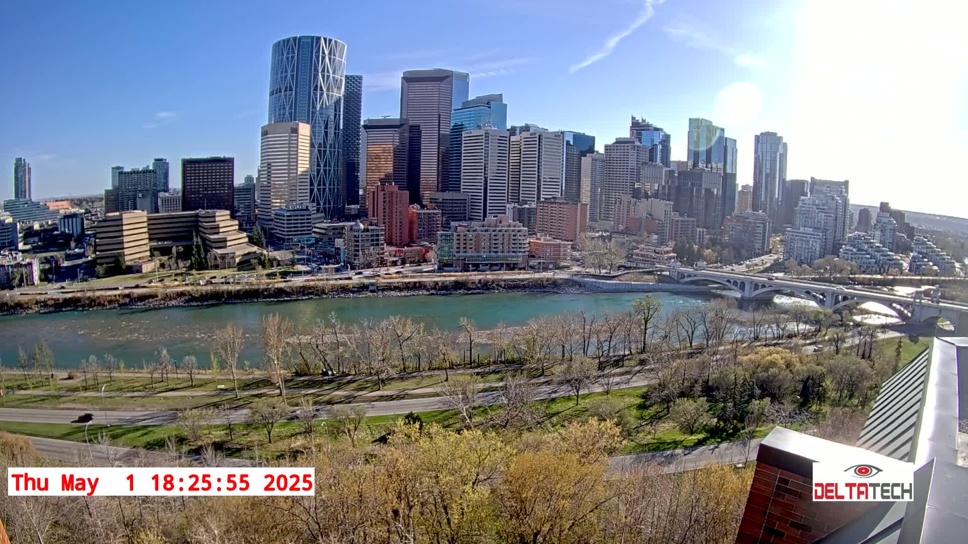 A sunny cityscape with numerous skyscrapers is viewed from across a river on a bright, clear day, with trees showing early spring growth.