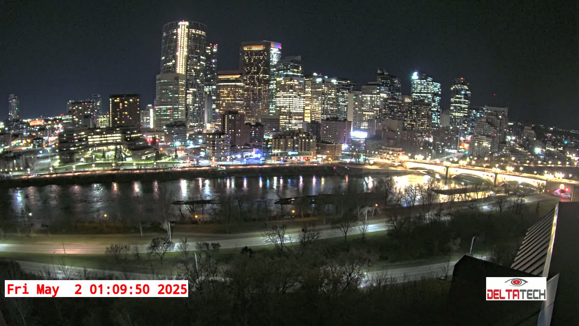 A brightly lit city skyline at night is reflected in a calm river, with a road curving along the riverbank.