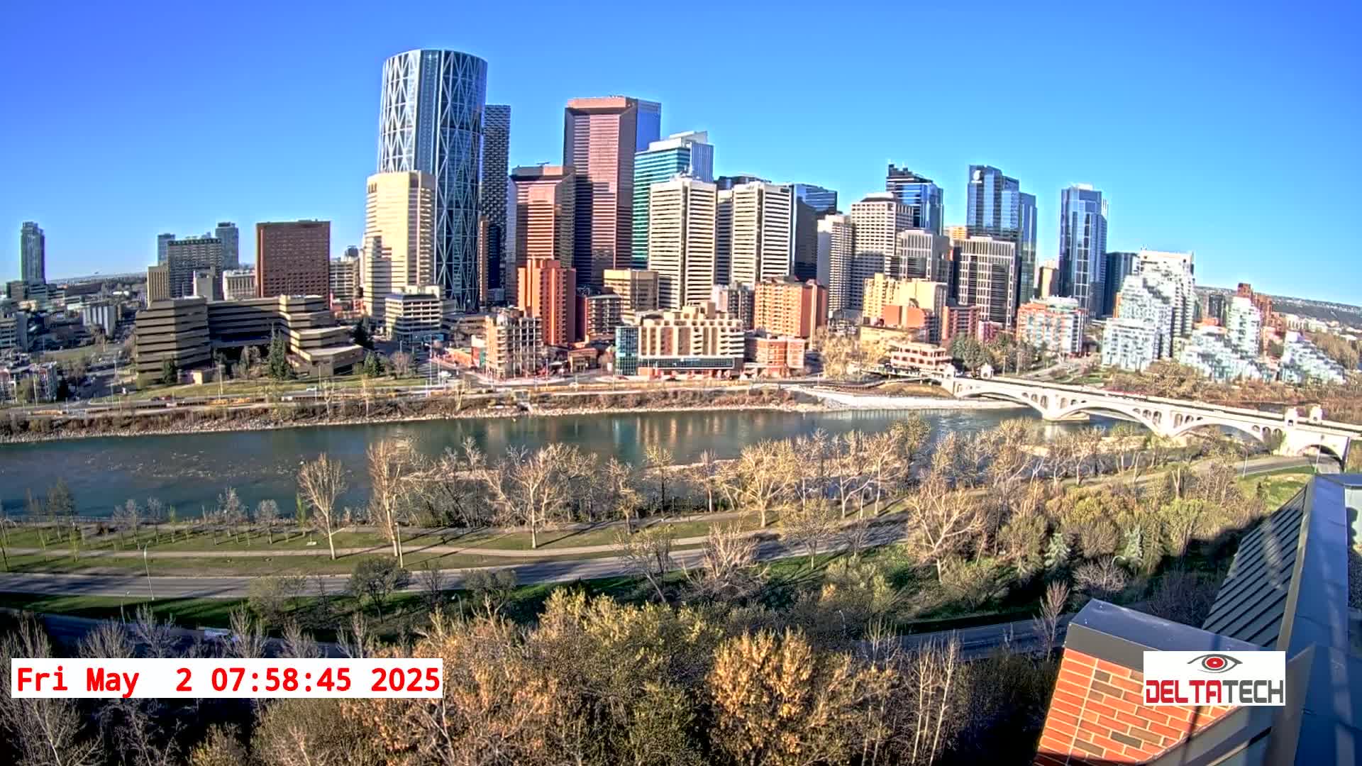 A cityscape with many tall buildings sits on the far bank of a wide river under a clear blue sky, with bare trees lining a pathway along the riverbank in the foreground.