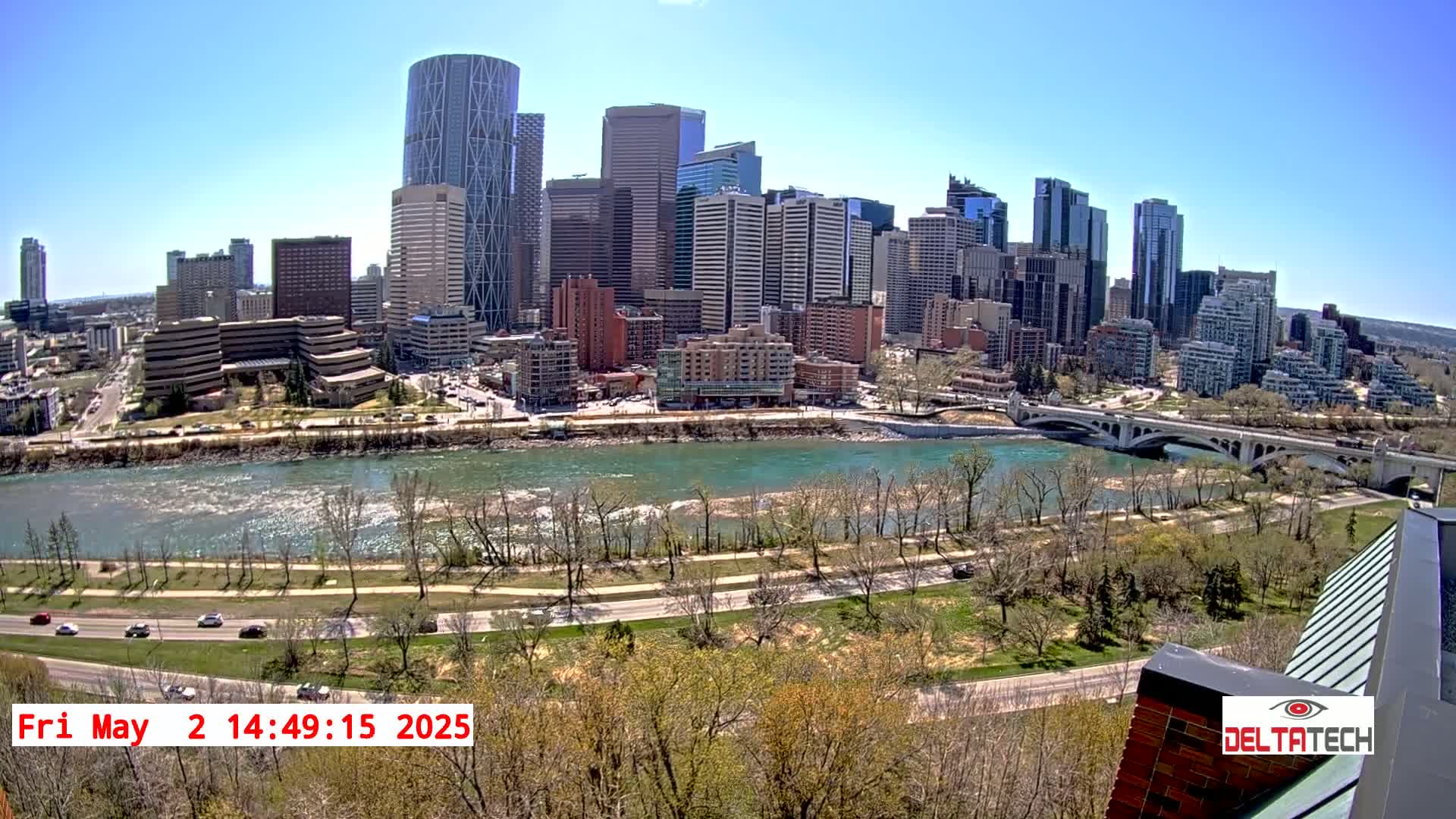 A city skyline with many tall buildings is seen across a wide river on a sunny day, with a bridge spanning the river and trees lining the riverbanks.