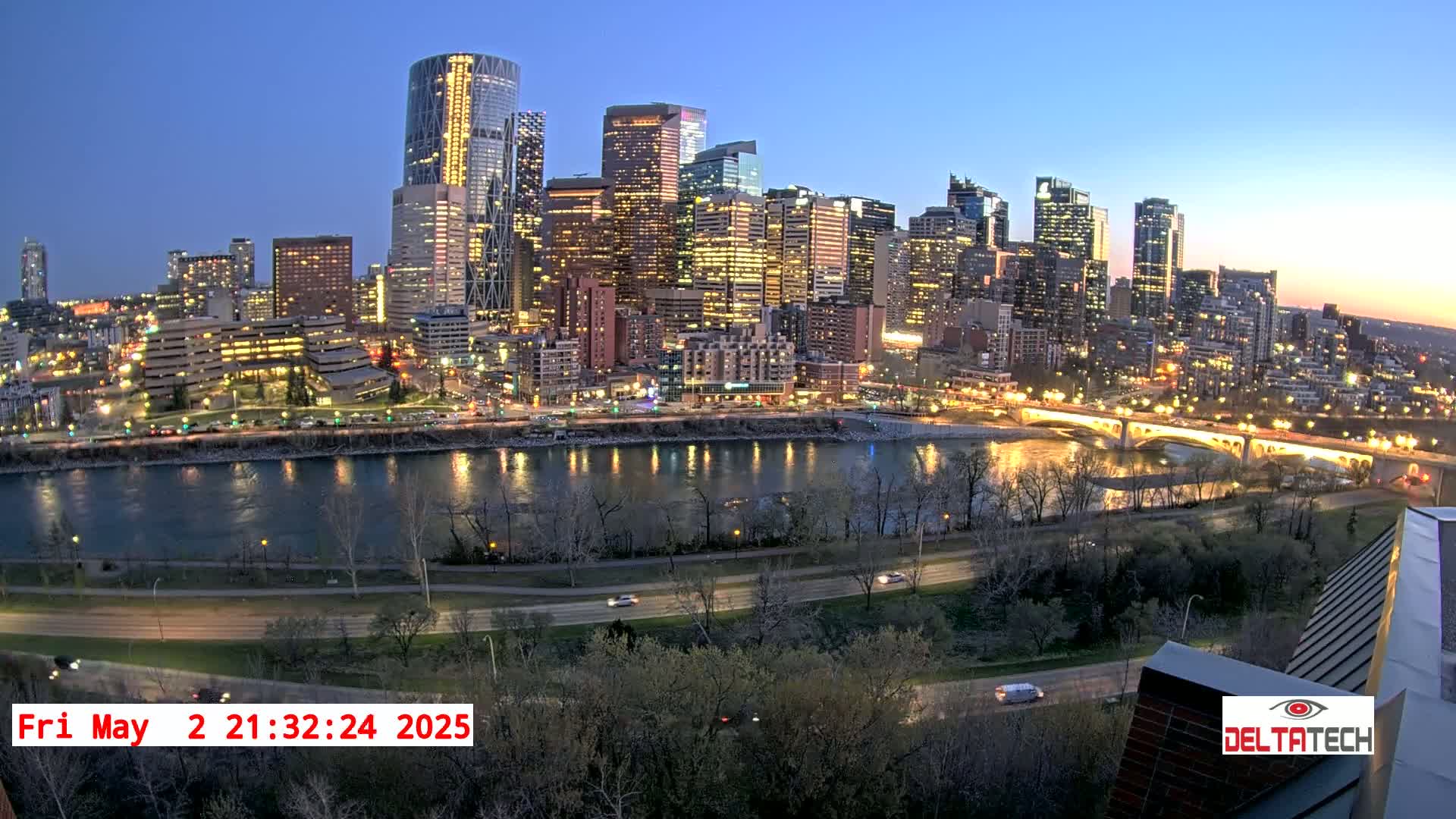 A city skyline illuminated at twilight, with a river reflecting the city lights, and a road winding through trees in the foreground.