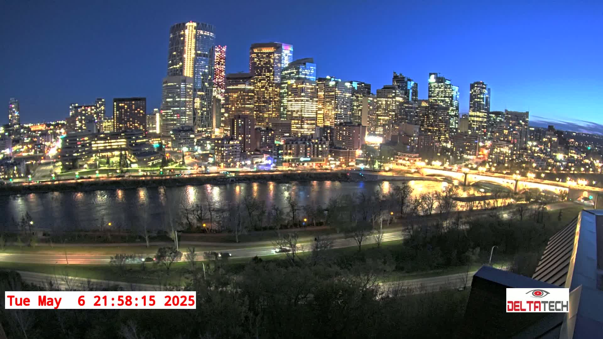 A brightly lit city skyline at night is situated across a calm river, bordered by trees and roads.