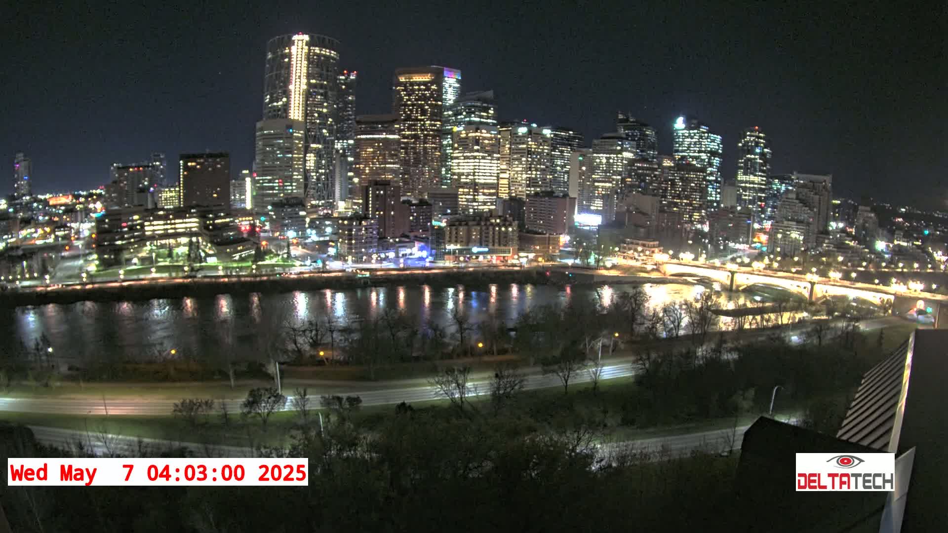 A brightly lit city skyline at night is seen across a dark river, with a roadway and trees in the foreground.