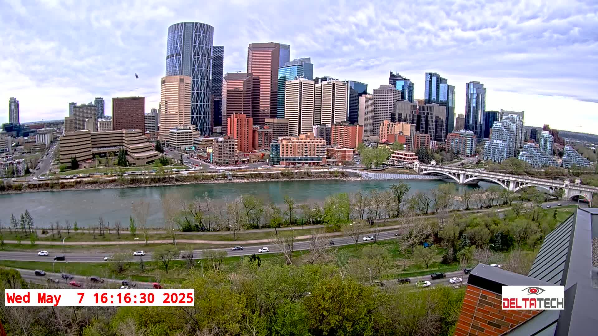 A city skyline with numerous high-rise buildings sits across a river, under a partly cloudy sky.