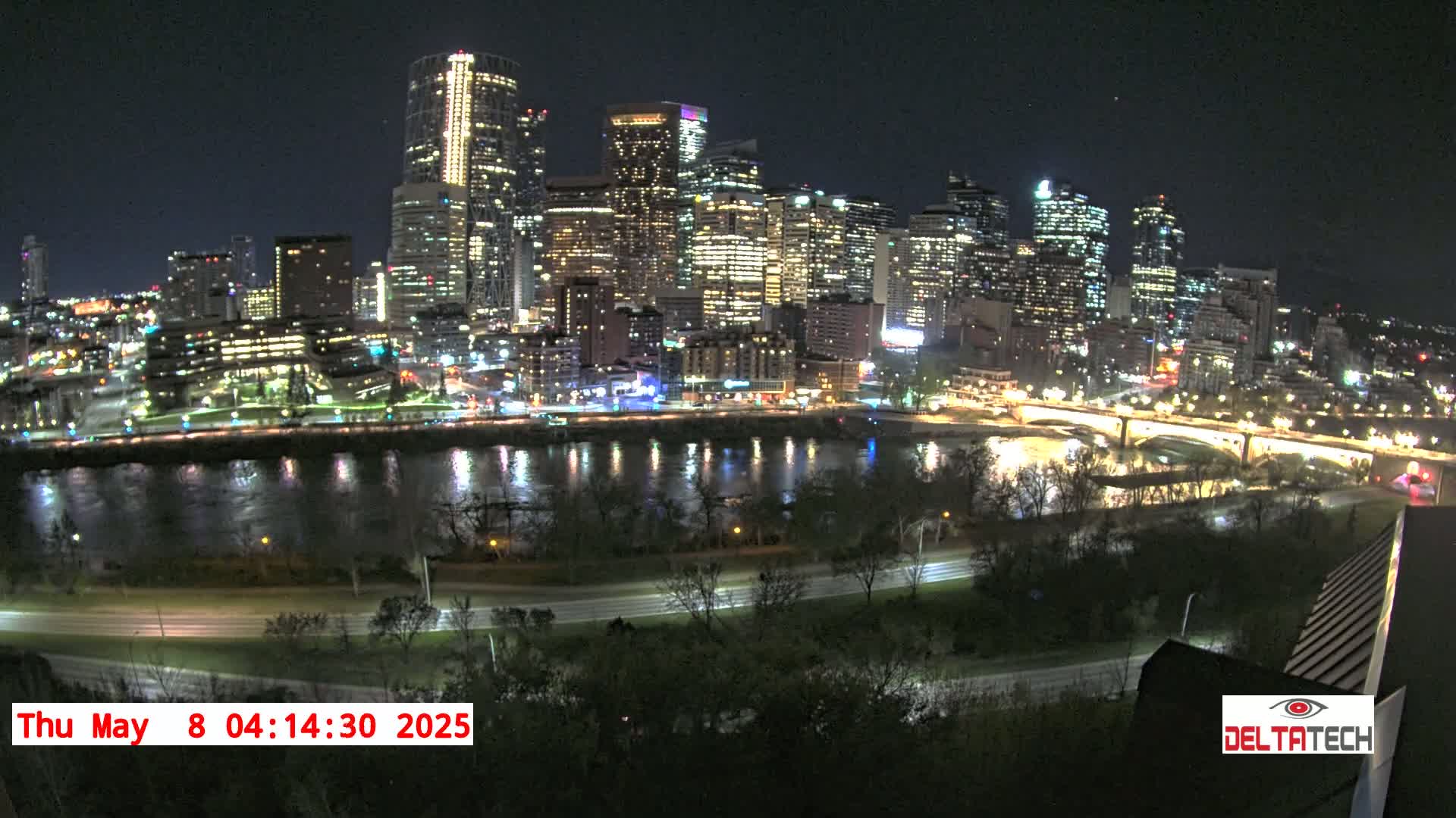 A nighttime view of a city skyline across a river, with illuminated buildings and bridges reflecting on calm water.