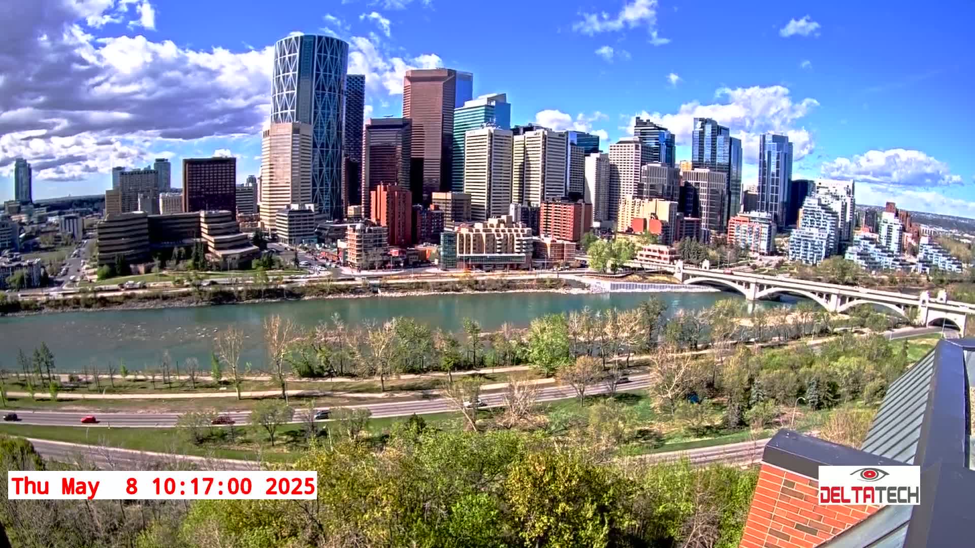 A city skyline with many tall buildings is seen across a river on a partly cloudy day, with green trees and a road along the riverbank.