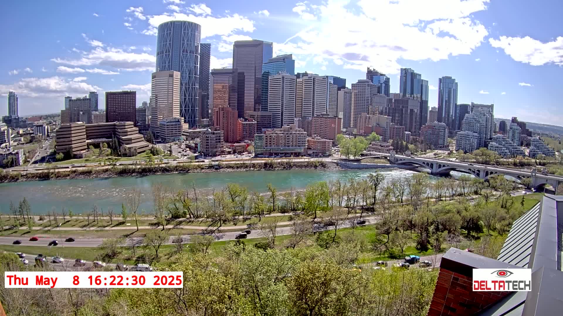 A city skyline with numerous skyscrapers is seen across a river under a partly cloudy, sunny sky.