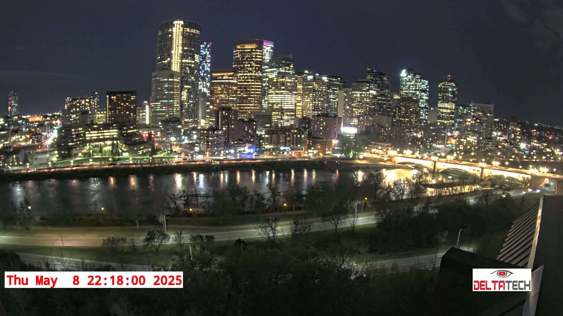 A brightly lit city skyline at night is reflected in a calm river, with trees and roads visible in the foreground under a clear sky.
