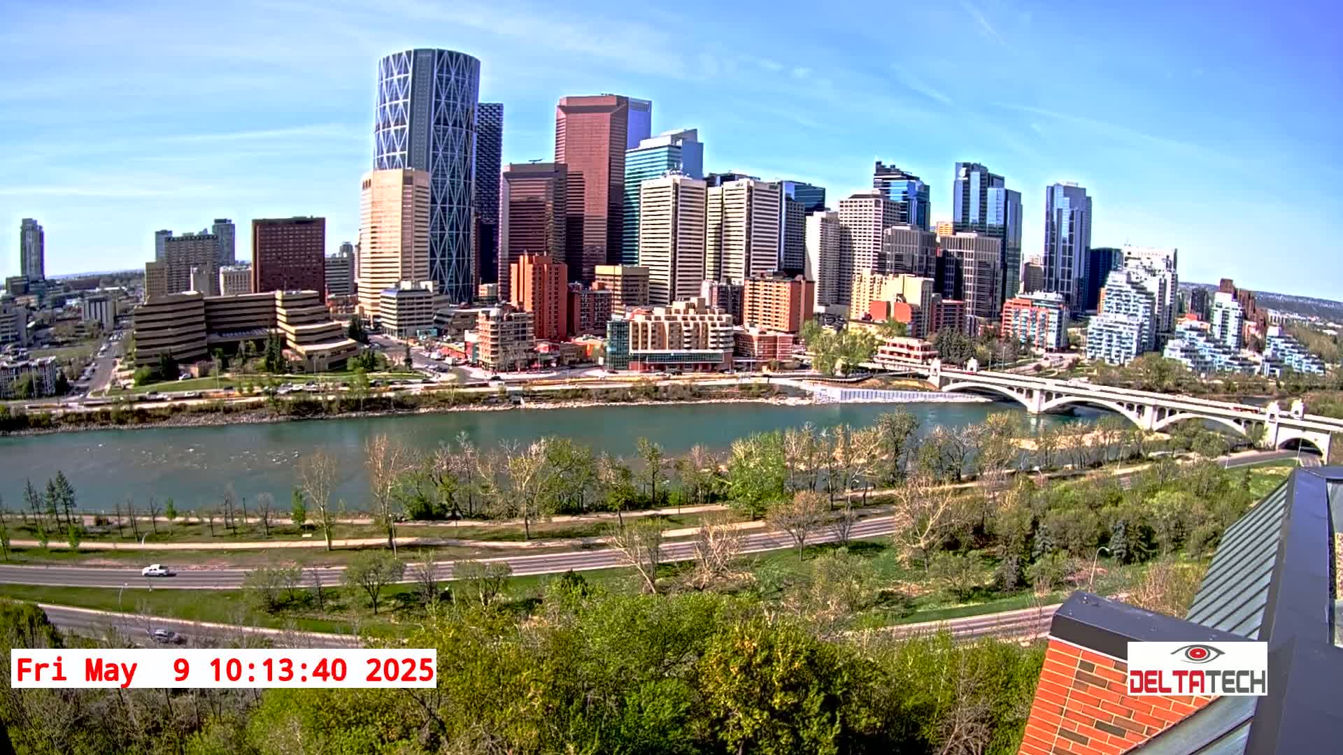 A city skyline with numerous high-rise buildings is seen across a river on a sunny day, with a bridge in the foreground and green vegetation lining the riverbanks.