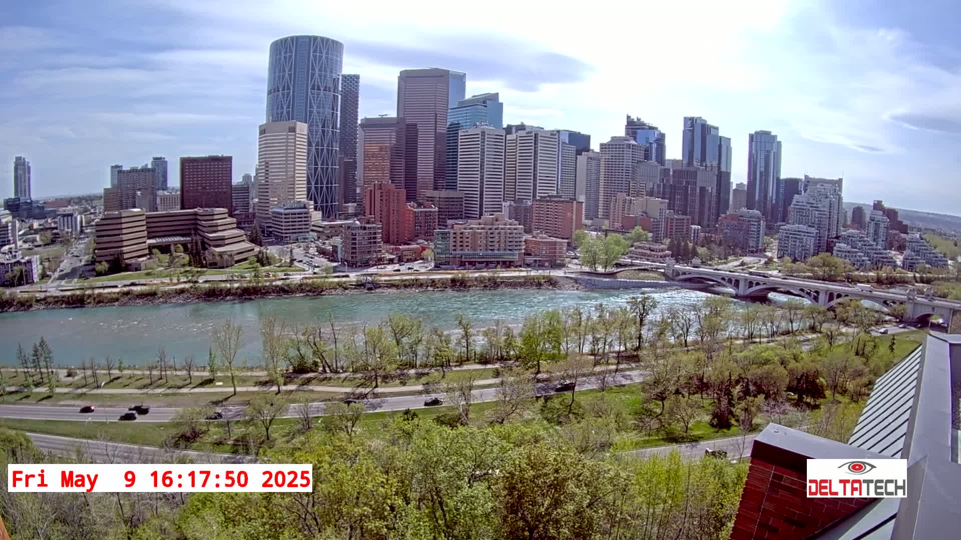 A city skyline with numerous skyscrapers and mid-rise buildings sits across a wide river under a partly cloudy sky, with a bridge visible in the foreground.