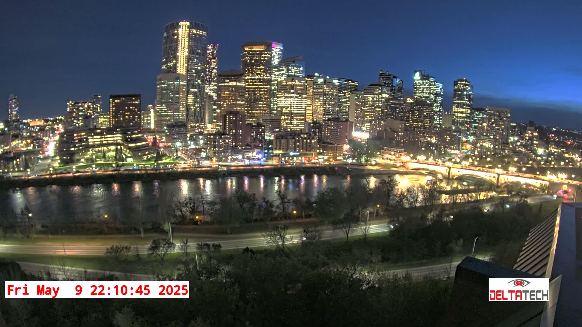 A brightly lit city skyline at night is reflected in a calm river, with a road and trees in the foreground under a dark blue, twilight sky.