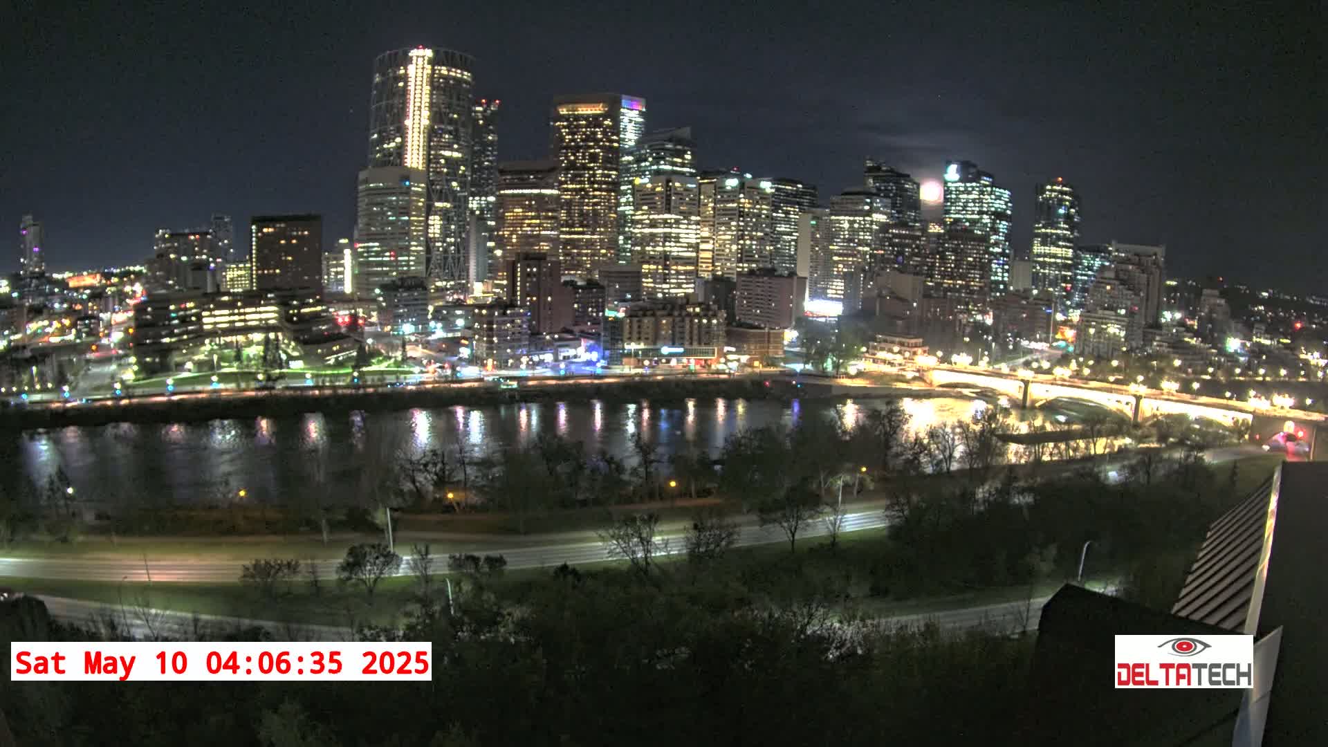 A brightly lit city skyline at night is reflected in a calm river, under a mostly clear sky with a visible moon.