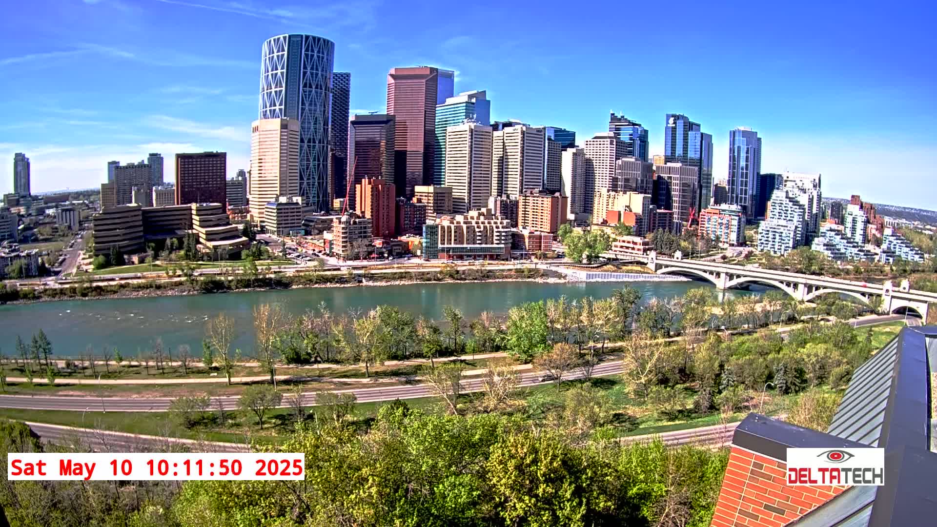 A city skyline with numerous skyscrapers and high-rise buildings is situated across a wide river under a clear, sunny sky.