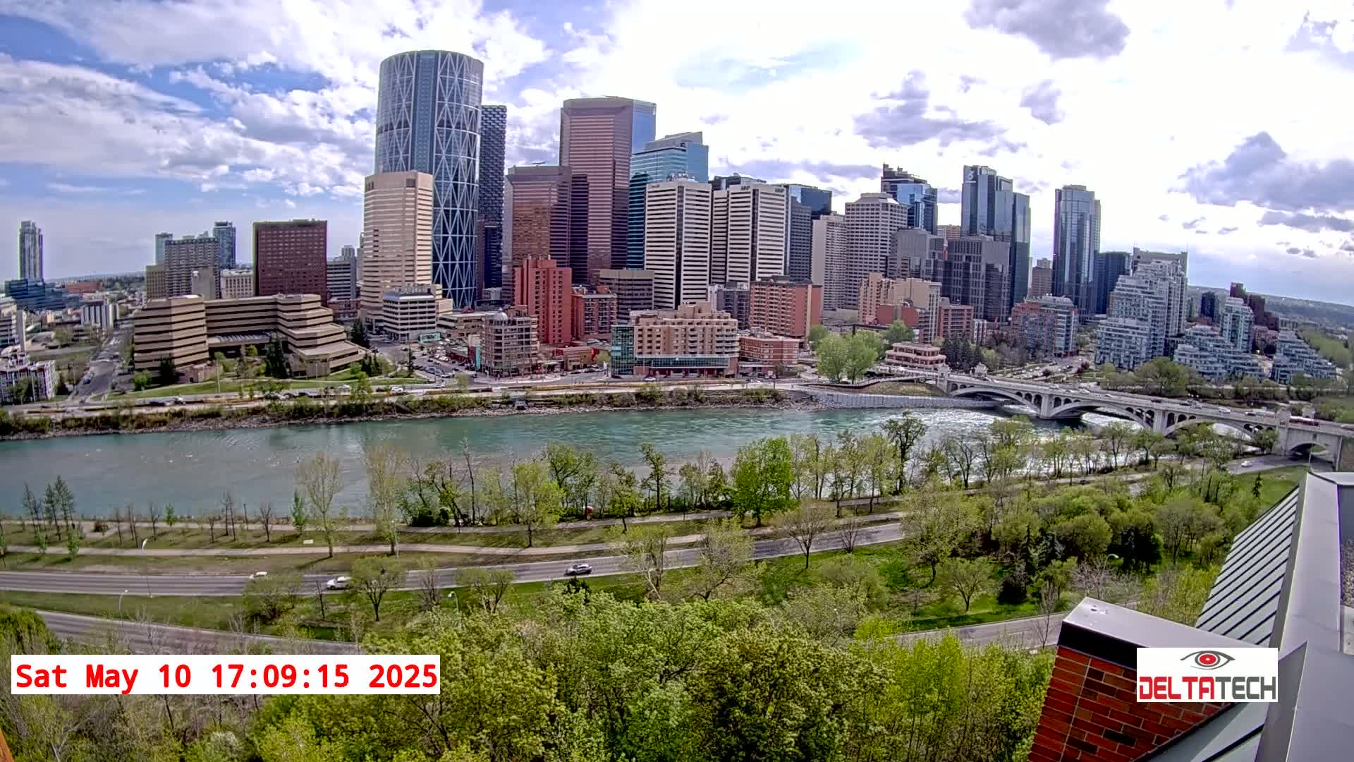 A city skyline with numerous skyscrapers sits across a river with a bridge under partly cloudy skies.