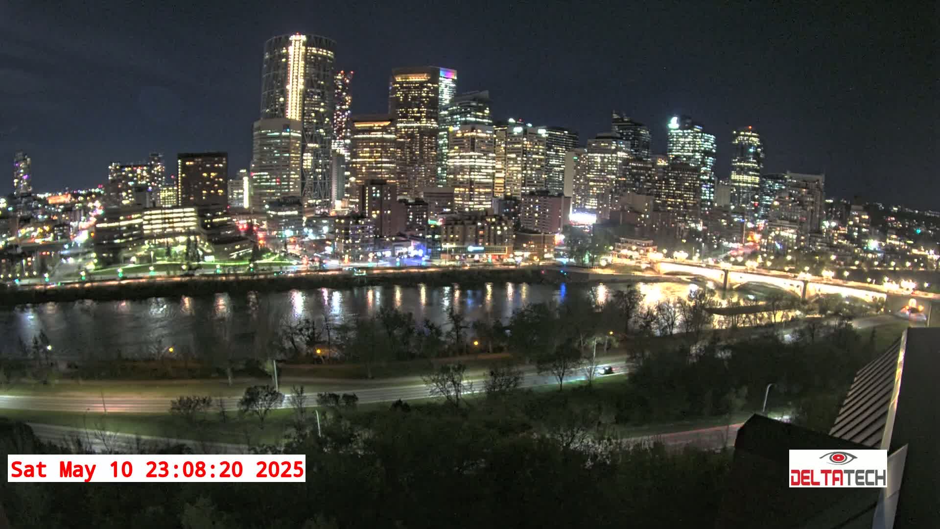 A nighttime view of a city skyline illuminated across a calm river, with trees and roads in the foreground under a clear sky.