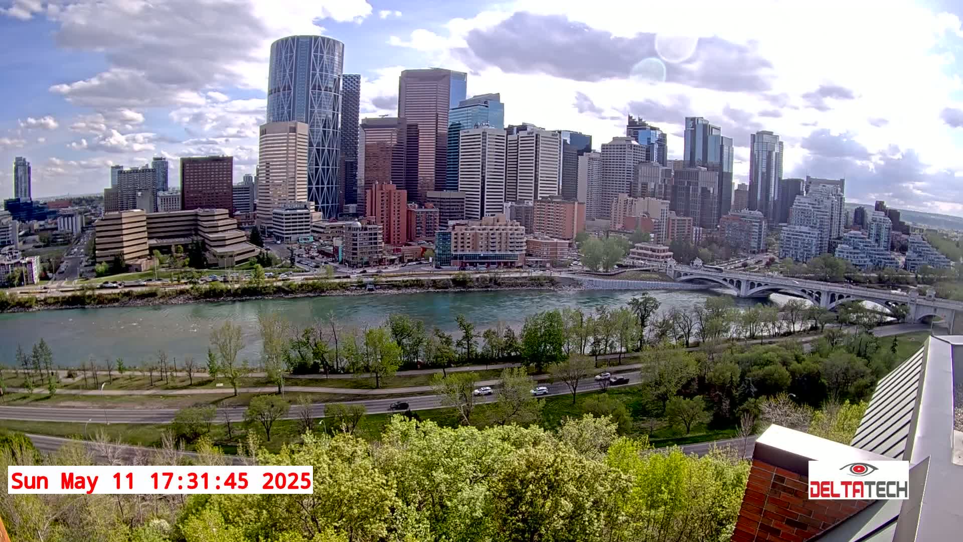 A city skyline with numerous skyscrapers sits across a wide river under a partly cloudy sky, with green trees and a road visible in the foreground.