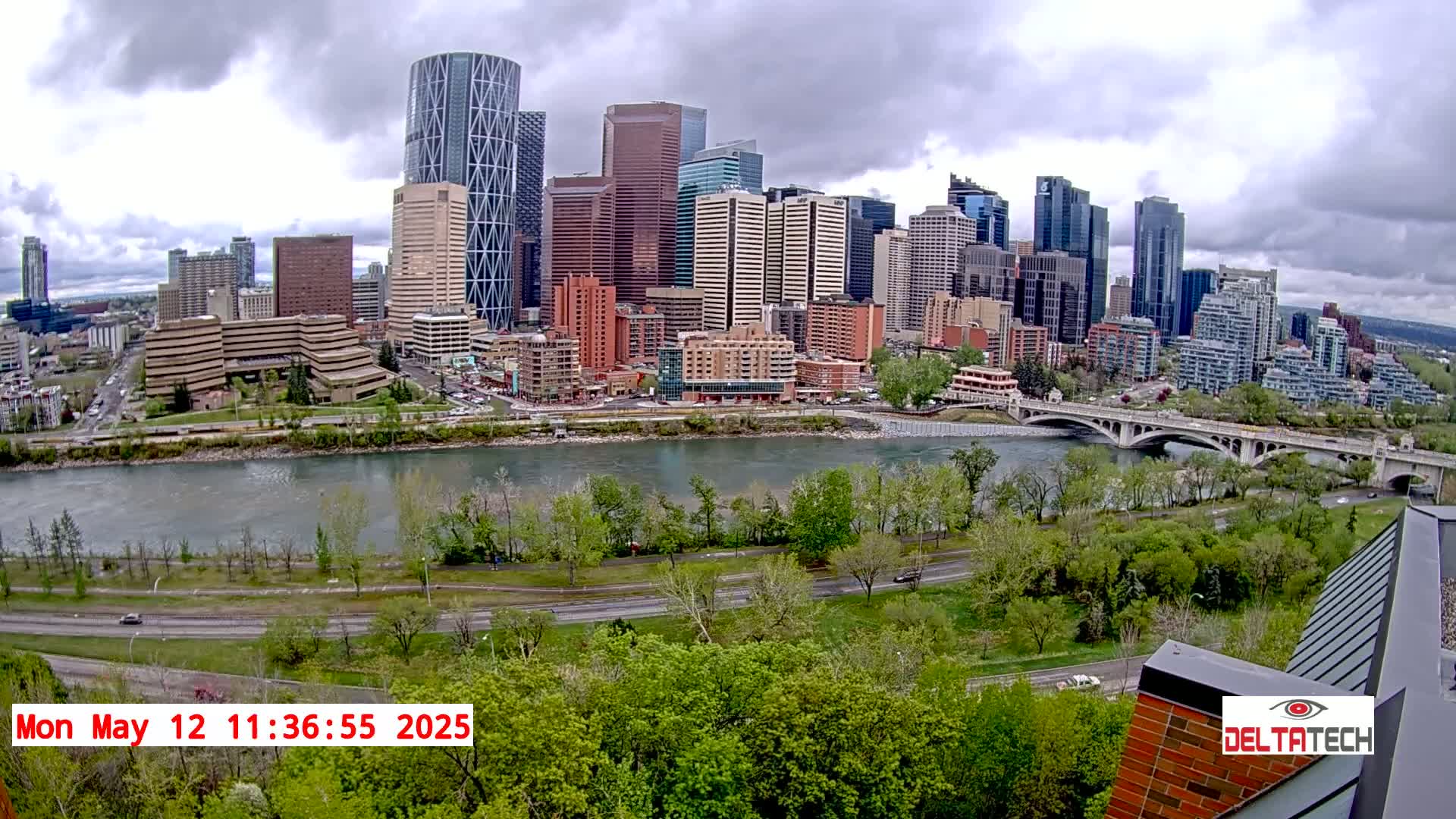 A city skyline with numerous skyscrapers is situated across a wide river from a green park, under a cloudy sky.