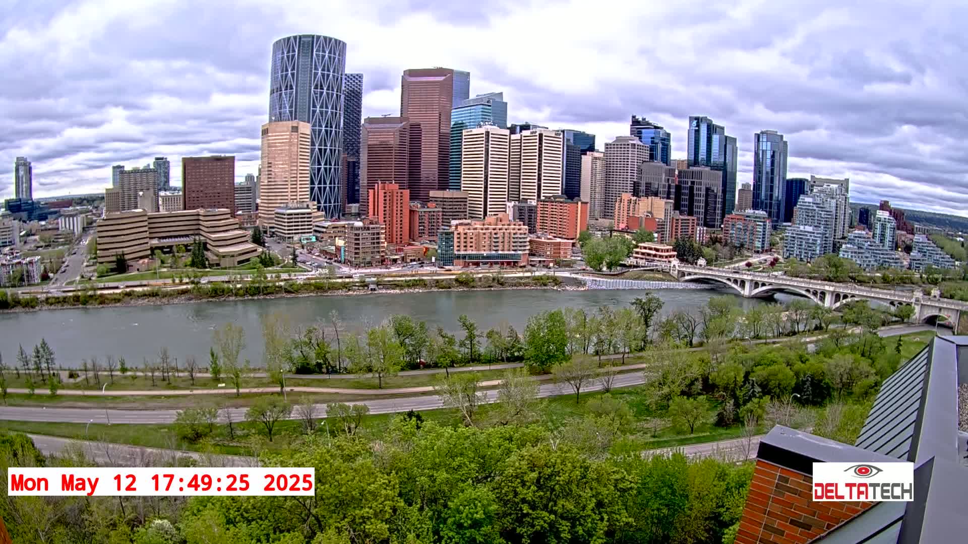 A city skyline with many tall buildings is viewed from across a river under a partly cloudy sky, with a bridge and lush green trees along the riverbank.