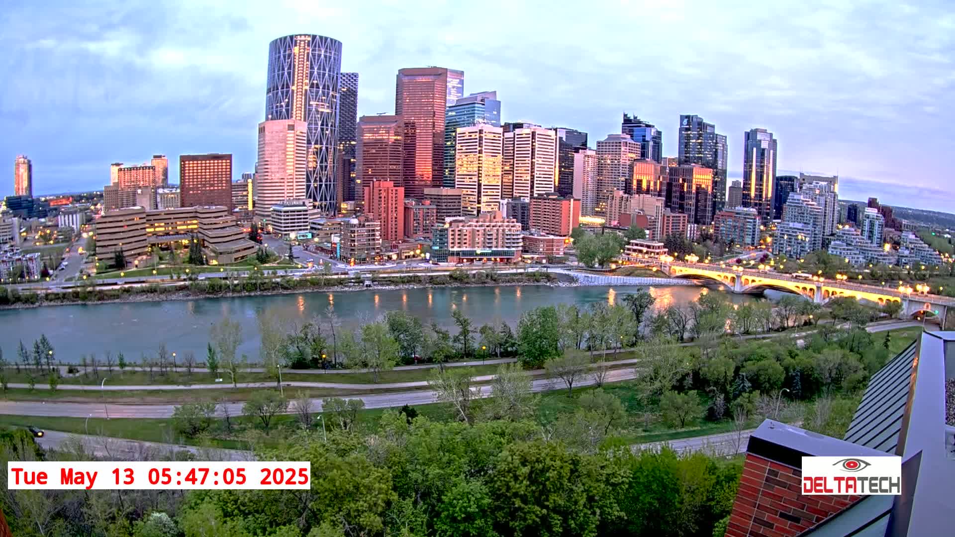 A city skyline with numerous skyscrapers is seen across a river at dusk, with trees and a roadway in the foreground.