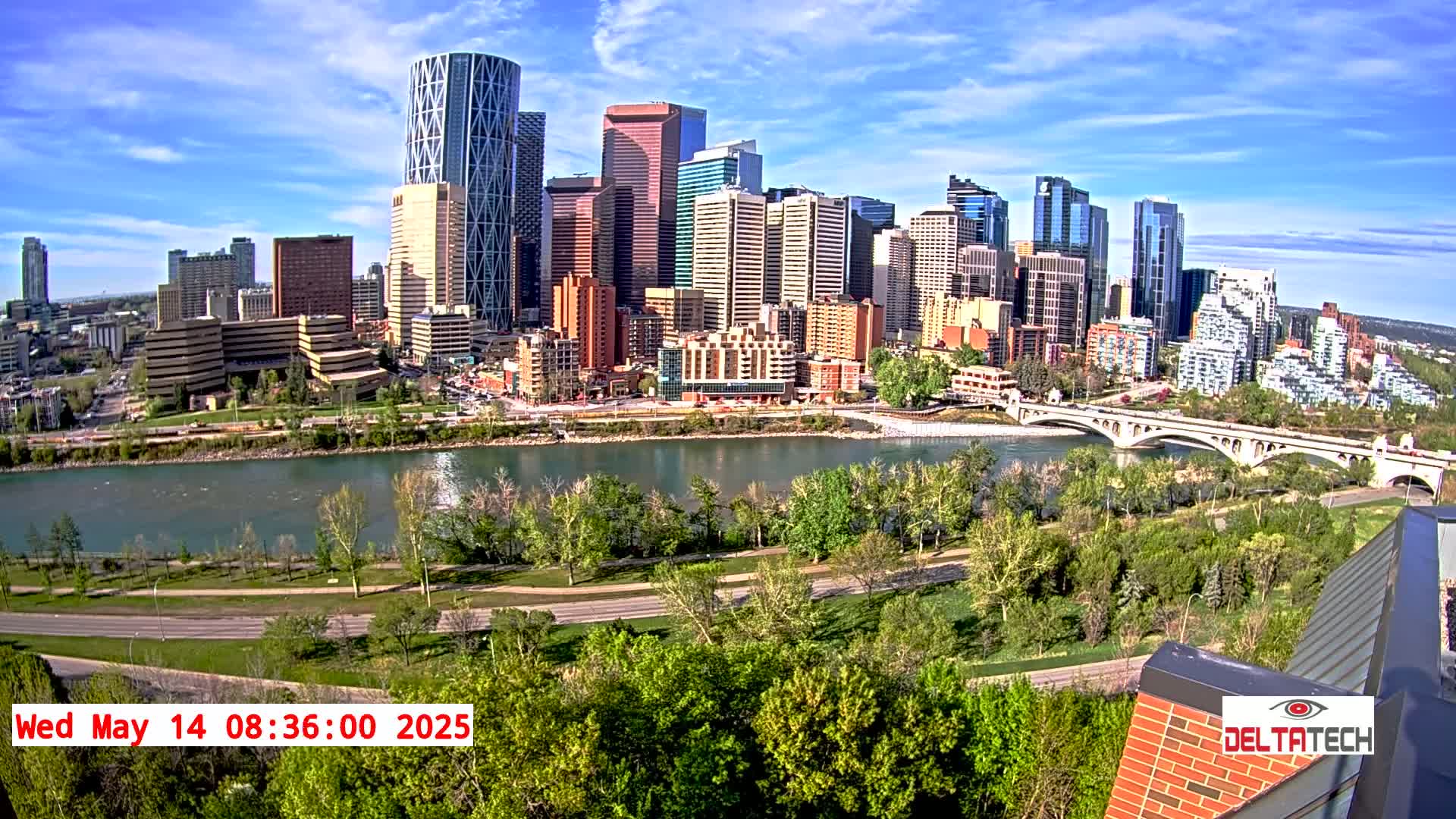 A city skyline with many tall buildings is seen across a river on a partly sunny day, with green trees lining the riverbanks and roads visible along the shore.