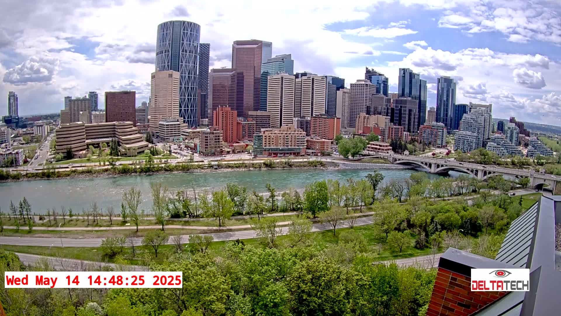 A city skyline with numerous skyscrapers is visible across a river, under partly cloudy skies, with lush green trees lining a roadway along the riverbank.