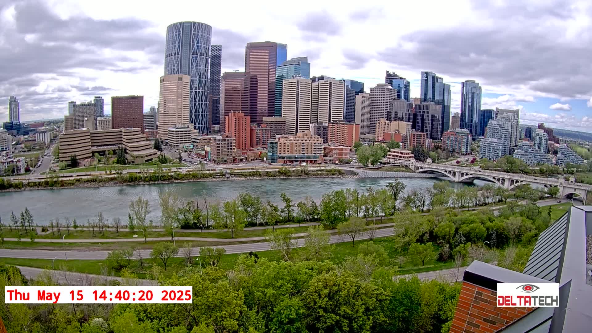 A city skyline with numerous skyscrapers is visible across a wide river, under partly cloudy skies, with lush green trees lining the riverbank and a bridge in the foreground.