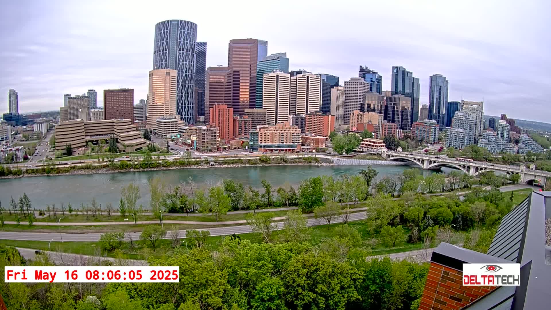 A city skyline with many tall buildings is viewed from across a river on an overcast day, with green trees lining the riverbank and a road curving alongside it.