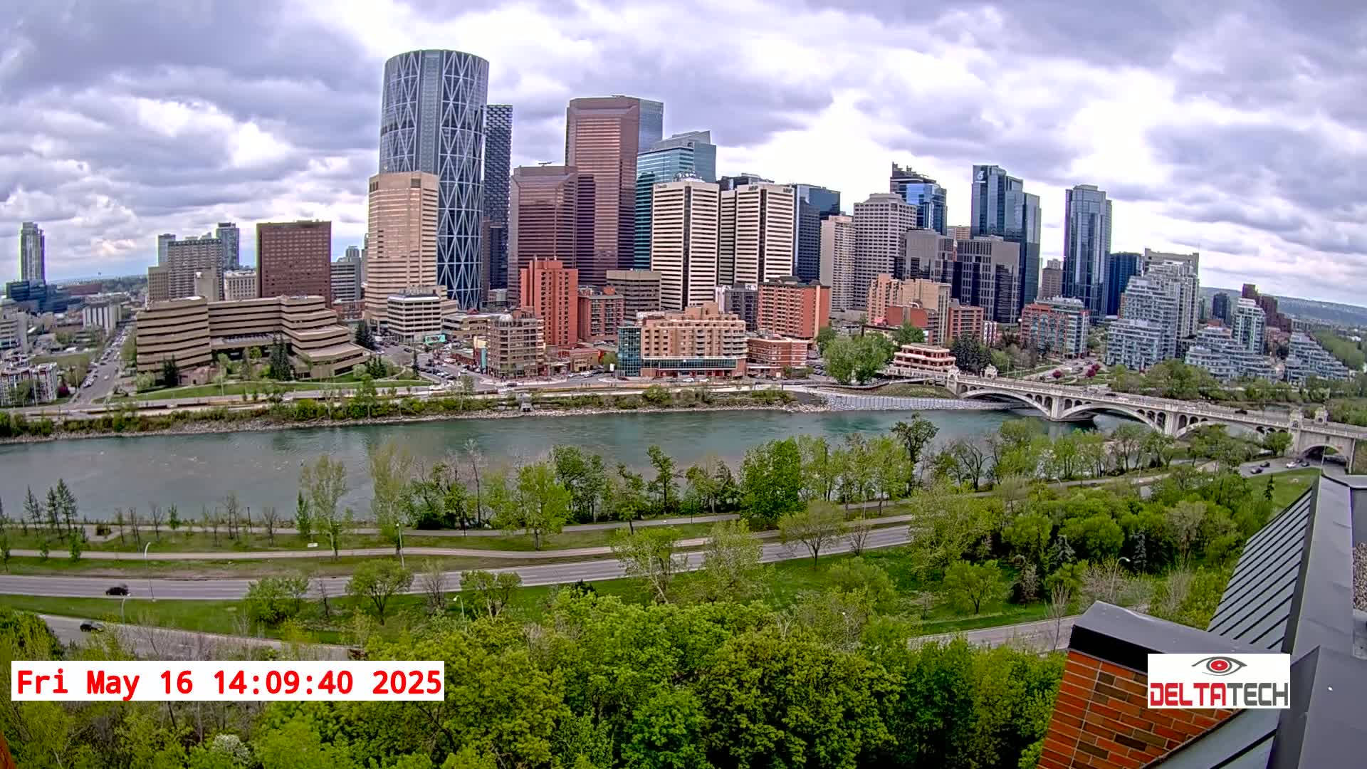 A city skyline with numerous skyscrapers and high-rise buildings sits across a river from a park with lush green trees under a partly cloudy sky.