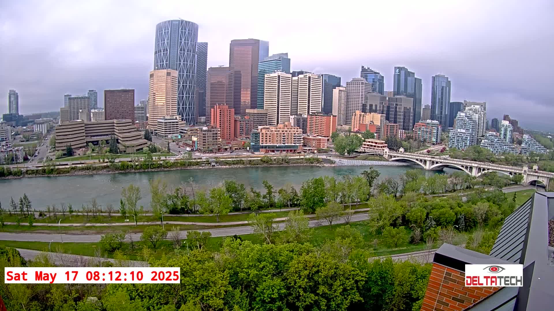 A city skyline with numerous skyscrapers and high-rise buildings sits across a river under an overcast sky, with lush green trees and a bridge in the foreground.