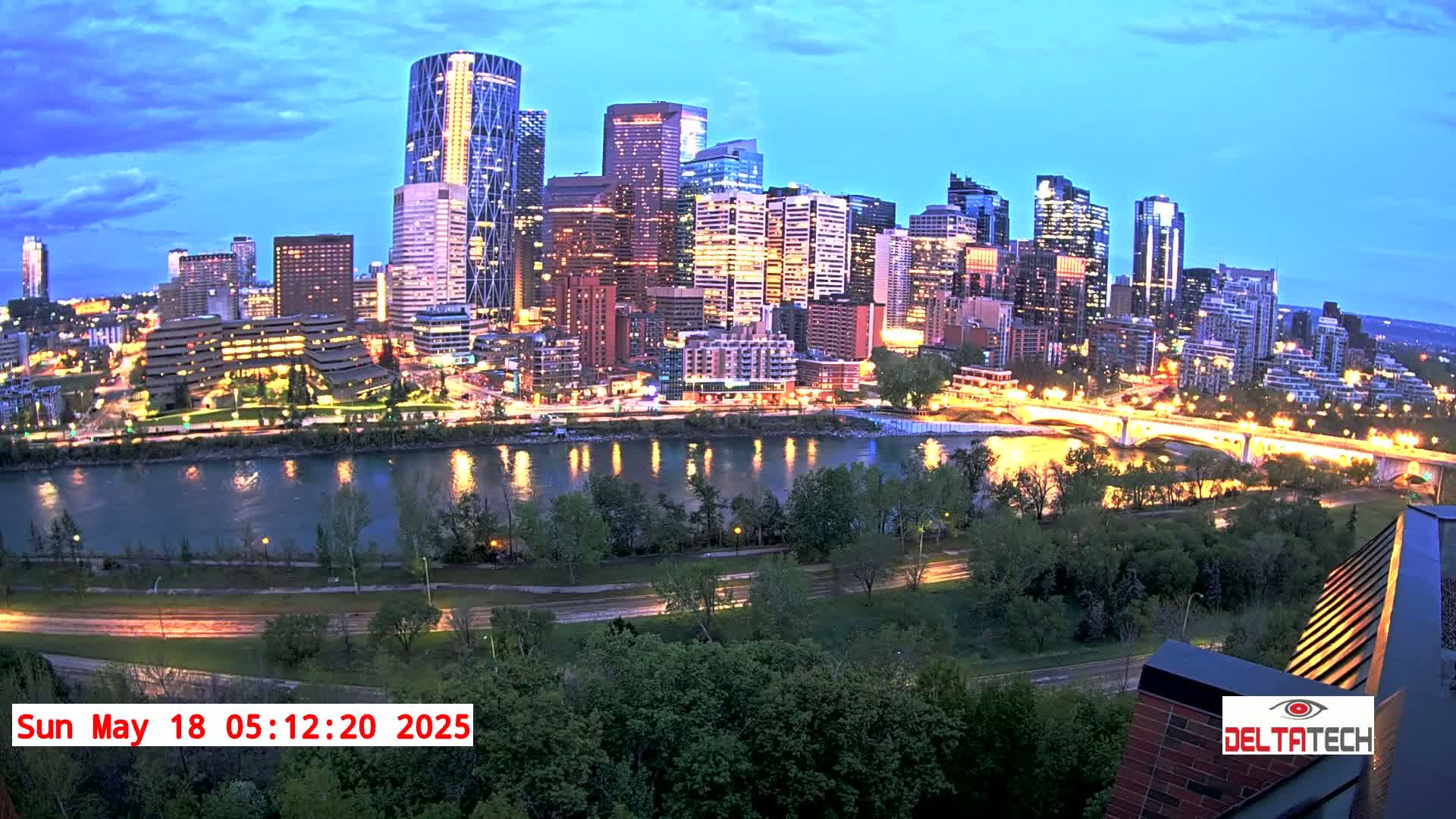 A city skyline at twilight, illuminated with city lights, sits beside a river and is surrounded by lush green trees under a partly cloudy sky.