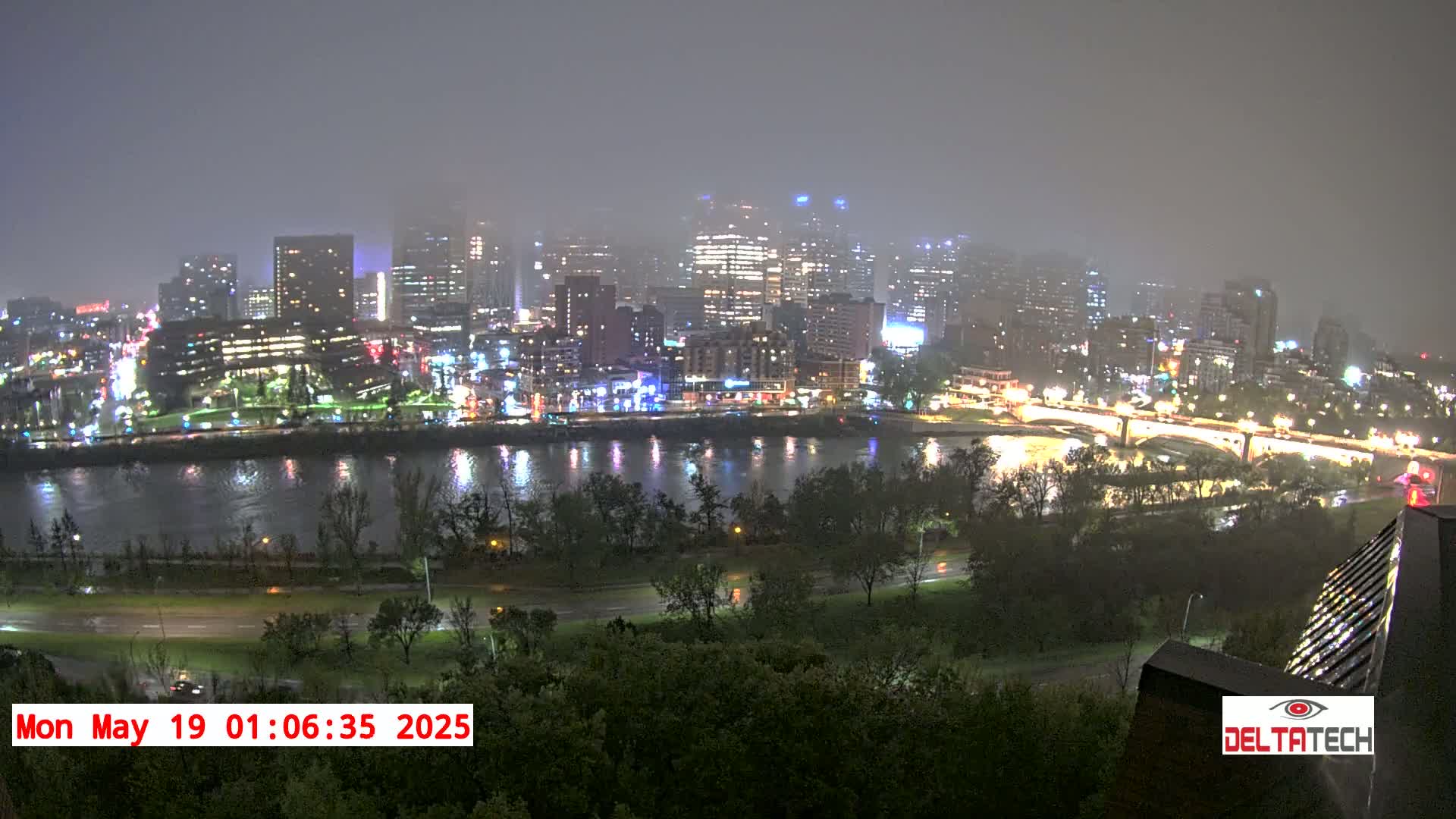 A nighttime cityscape with illuminated buildings and a river reflecting the city lights is seen through a foggy or misty atmosphere.